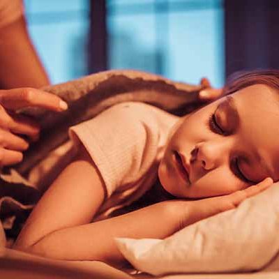 Child sleeping on her side with a mother's hands tucking the blanket around her shoulders